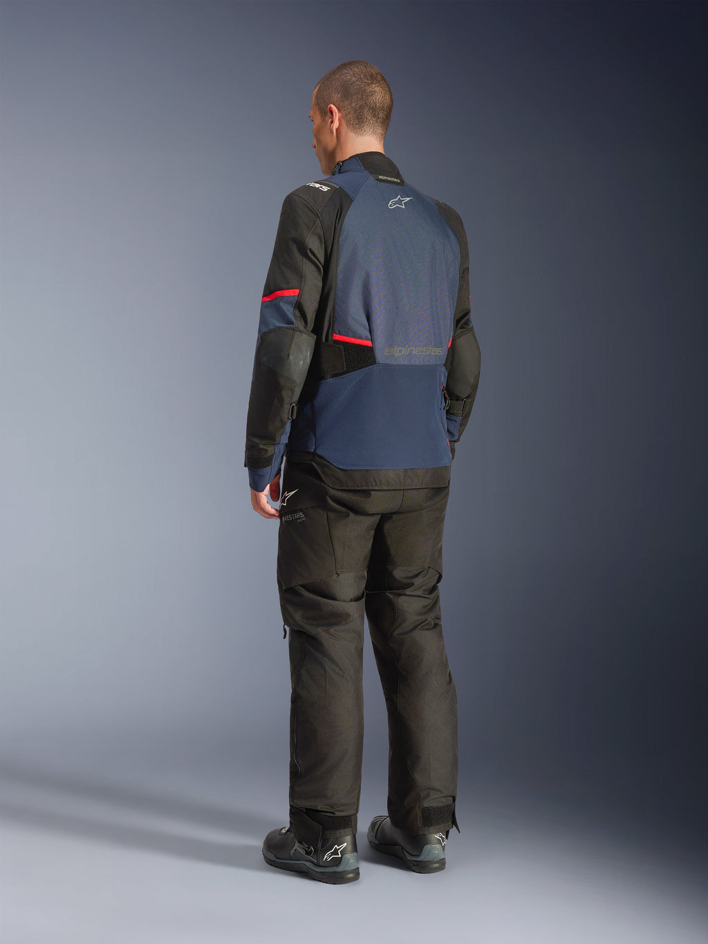 Alpinestars Andes V4 Drystar® Jacket, giacca da moto in Night Navy e Mood Indigo con pannelli neri e accenti rossi, vista posteriore di tre quarti, indossata con pantaloni da moto adventure neri e stivali, costruzione in tessuto tecnico con logo Alpinestars sulla schiena, orlo posteriore allungato.