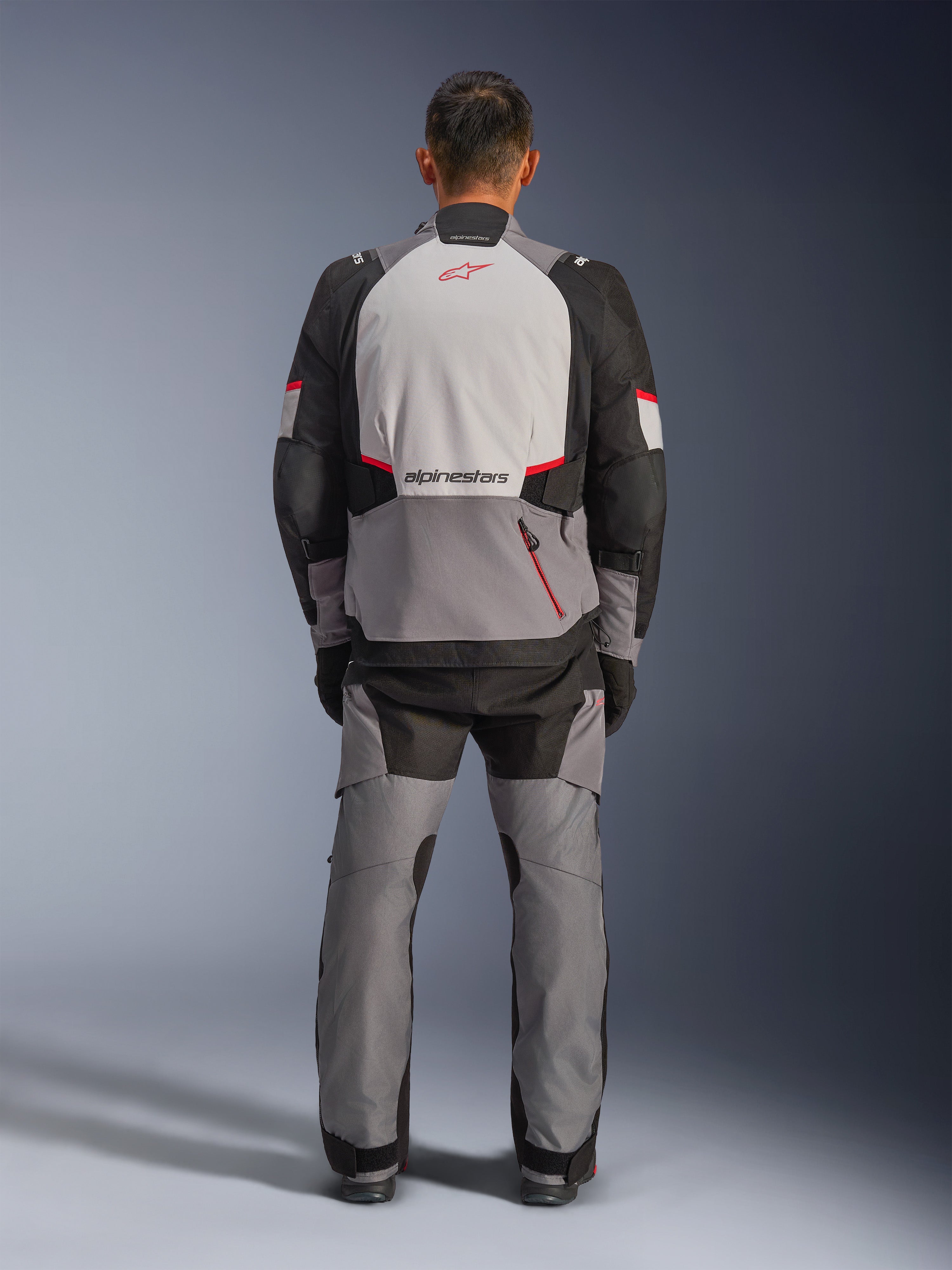Giacca Alpinestars Andes V4 Drystar®, giacca da moto adventure, grigio scuro, grigio ghiaccio, nero e rosso brillante, vista posteriore di un pilota che indossa pantaloni coordinati, con un ampio pannello posteriore grigio ghiaccio e logo rosso, sezioni rinforzate sulle spalle nere e cinghie regolabili in vita per il touring in ogni condizione atmosferica.