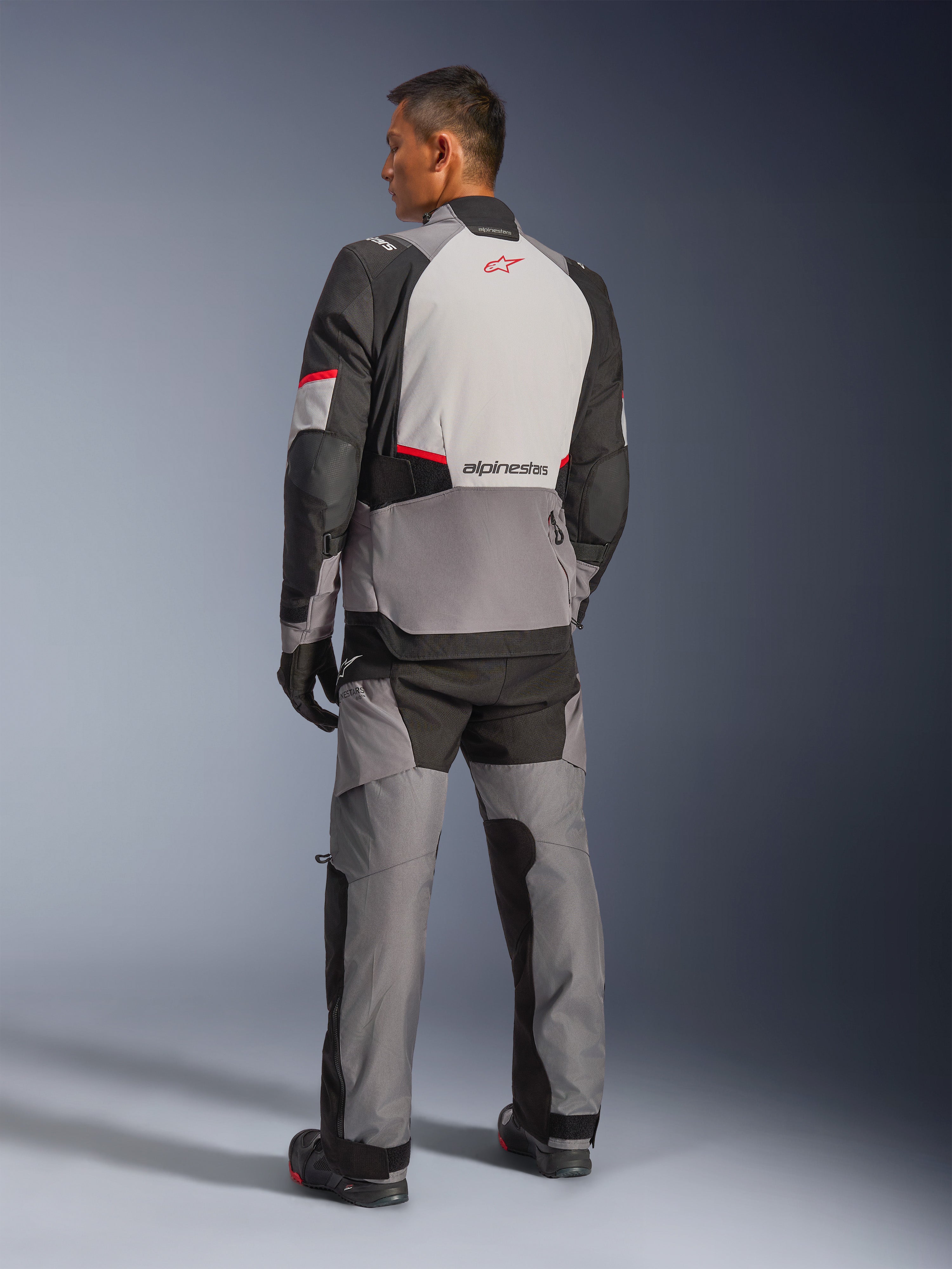 Giacca Alpinestars Andes V4 Drystar®, grigio scuro e grigio ghiaccio con accenti nero e rosso brillante, vista posteriore che mostra il pannello grigio chiaro con logo della stella rossa, abbinata a pantaloni adventure coordinati e guanti neri, abbigliamento tecnico da moto per ogni condizione atmosferica.