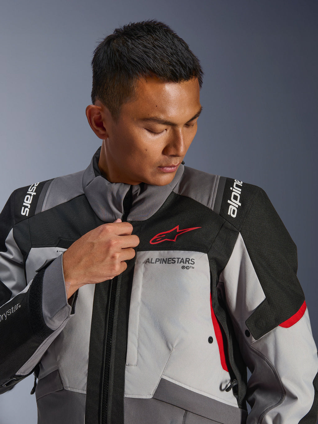 Modello che indossa la giacca Alpinestars Andes V4 Drystar®, grigio scuro e grigio ghiaccio con accenti neri e rosso acceso, regola la cerniera frontale, con logo Astars rosso e marchio Drystar®, stile adventure touring