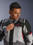 Modello che indossa la giacca Alpinestars Andes V4 Drystar®, grigio scuro e grigio ghiaccio con accenti neri e rosso acceso, regola la cerniera frontale, con logo Astars rosso e marchio Drystar®, stile adventure touring