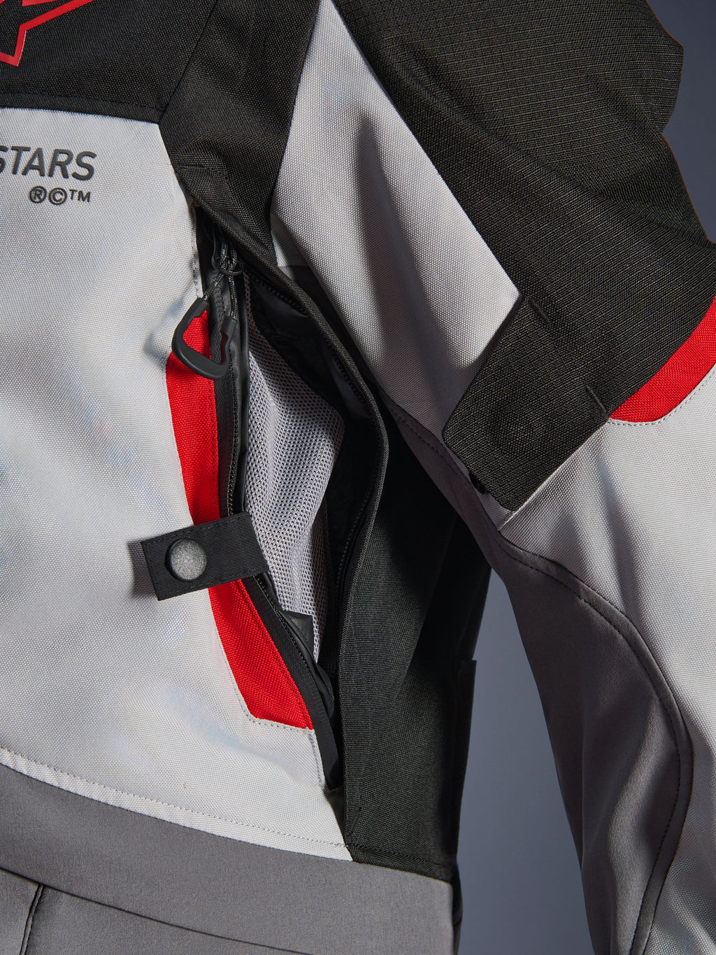 Dettaglio ravvicinato della giacca Alpinestars Andes V4 Drystar® in grigio scuro, grigio ghiaccio e rosso acceso, che mette in risalto una presa d'aria sul petto con cerniera, fodera in rete e un cinturino di regolazione con bottone a pressione.