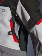 Dettaglio ravvicinato della giacca Alpinestars Andes V4 Drystar® in grigio scuro, grigio ghiaccio e rosso acceso, che mette in risalto una presa d'aria sul petto con cerniera, fodera in rete e un cinturino di regolazione con bottone a pressione.