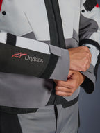 Dettaglio ravvicinato della manica della giacca Alpinestars Andes V4 Drystar®, tessuto grigio e nero, con logo Drystar rosso e bianco sull'avambraccio, chiusura del polso regolabile in velcro, abbigliamento da moto impermeabile ad alte prestazioni