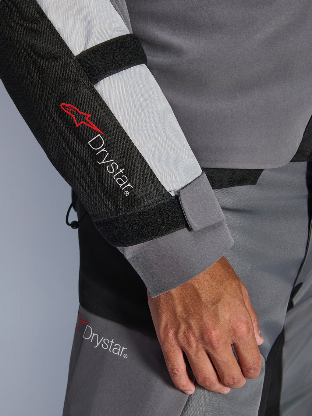 Primo piano della manica della giacca Alpinestars Andes V4 Drystar®, grigio scuro e grigio ghiaccio con dettagli neri, con logo Drystar® e cinghie regolabili sull'avambraccio, parte di un completo da moto adventure in tessuto impermeabile