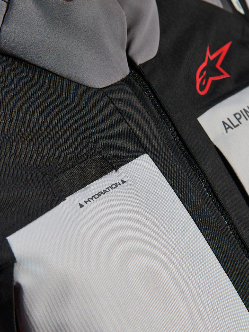 Giacca Alpinestars Andes V4 Drystar®, dettaglio ravvicinato del petto, pannelli grigio scuro e grigio ghiaccio con rinforzi in tessuto nero, logo Alpinestars rosso e passante per il tubo di idratazione con etichetta 'HYDRATION'