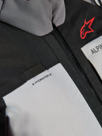 Giacca Alpinestars Andes V4 Drystar®, dettaglio ravvicinato del petto, pannelli grigio scuro e grigio ghiaccio con rinforzi in tessuto nero, logo Alpinestars rosso e passante per il tubo di idratazione con etichetta 'HYDRATION'