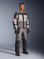 Modello con giacca Alpinestars Andes V4 Drystar® e pantaloni adventure coordinati, grigio scuro, grigio ghiaccio, nero e rosso acceso, abbinati a guanti e stivali da moto neri, abbigliamento tecnico per ogni condizione climatica con design moderno a più pannelli.