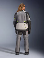 Giacca Alpinestars Andes V4 Drystar®, giacca da moto adventure, grigio scuro e grigio ghiaccio con accenti neri e giallo fluo, vista posteriore indossata con pantaloni coordinati e guanti neri, dotata di una grande tasca posteriore con cerniera gialla e pannelli rinforzati su spalle e gomiti
