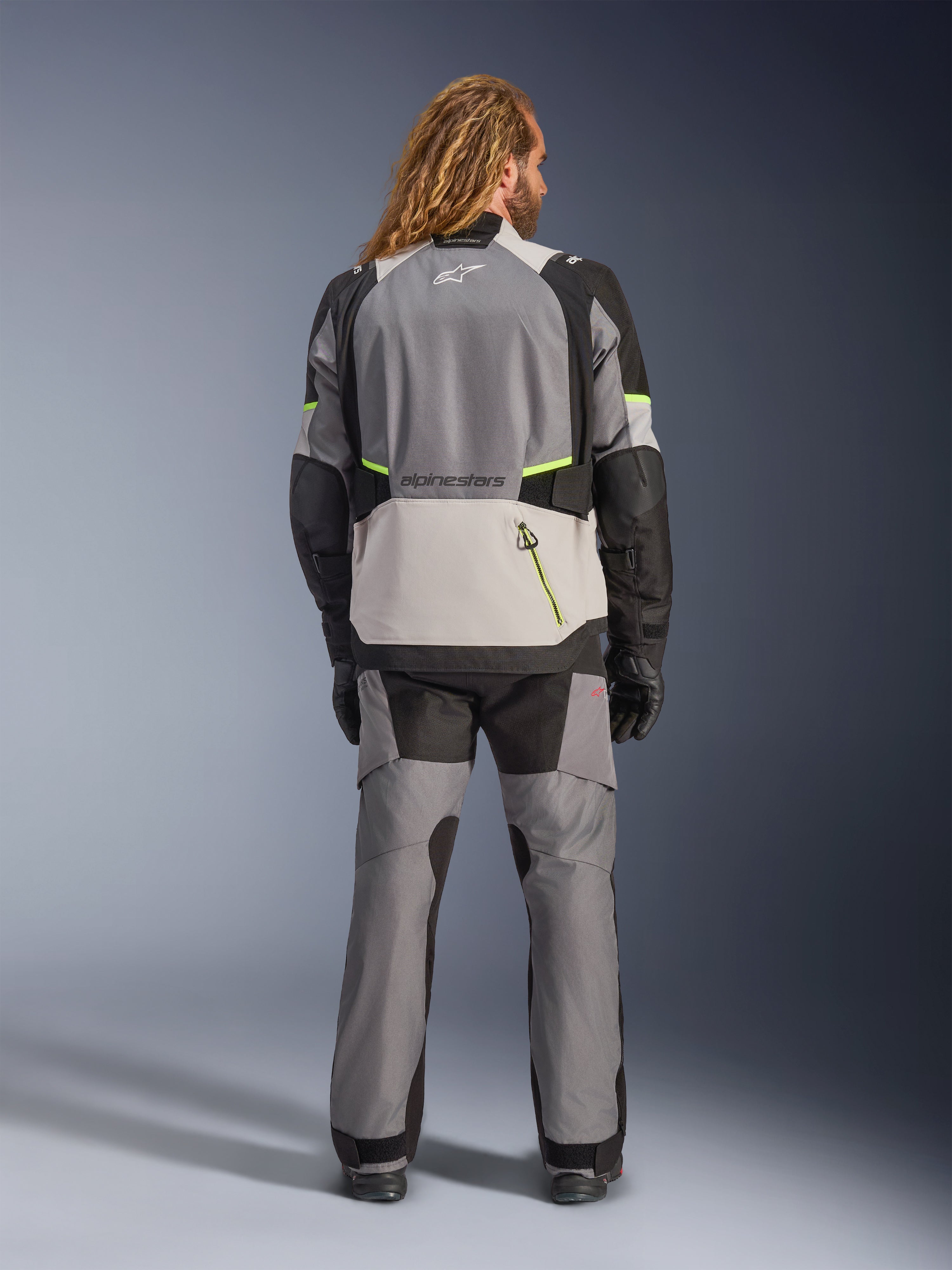 Giacca Alpinestars Andes V4 Drystar®, giacca da moto adventure, grigio scuro e grigio ghiaccio con accenti neri e giallo fluo, vista posteriore indossata con pantaloni coordinati e guanti neri, dotata di una grande tasca posteriore con cerniera gialla e pannelli rinforzati su spalle e gomiti