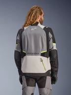 Giacca Alpinestars Andes V4 Drystar®, vista posteriore di giacca da moto adventure in grigio scuro, grigio ghiaccio e nero con accenti giallo fluo, con pannelli in tessuto rinforzato e loghi Alpinestars, indossata con pantaloni da moto coordinati e guanti neri