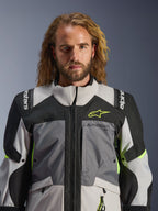 Giacca Alpinestars Andes V4 Drystar®, grigio scuro, grigio ghiaccio, nero e giallo fluo, indossata da un uomo con lunghi capelli mossi, caratterizzata da una costruzione tecnica multi-tessuto, loghi Alpinestars su petto e spalle, design a collo alto per la guida adventure