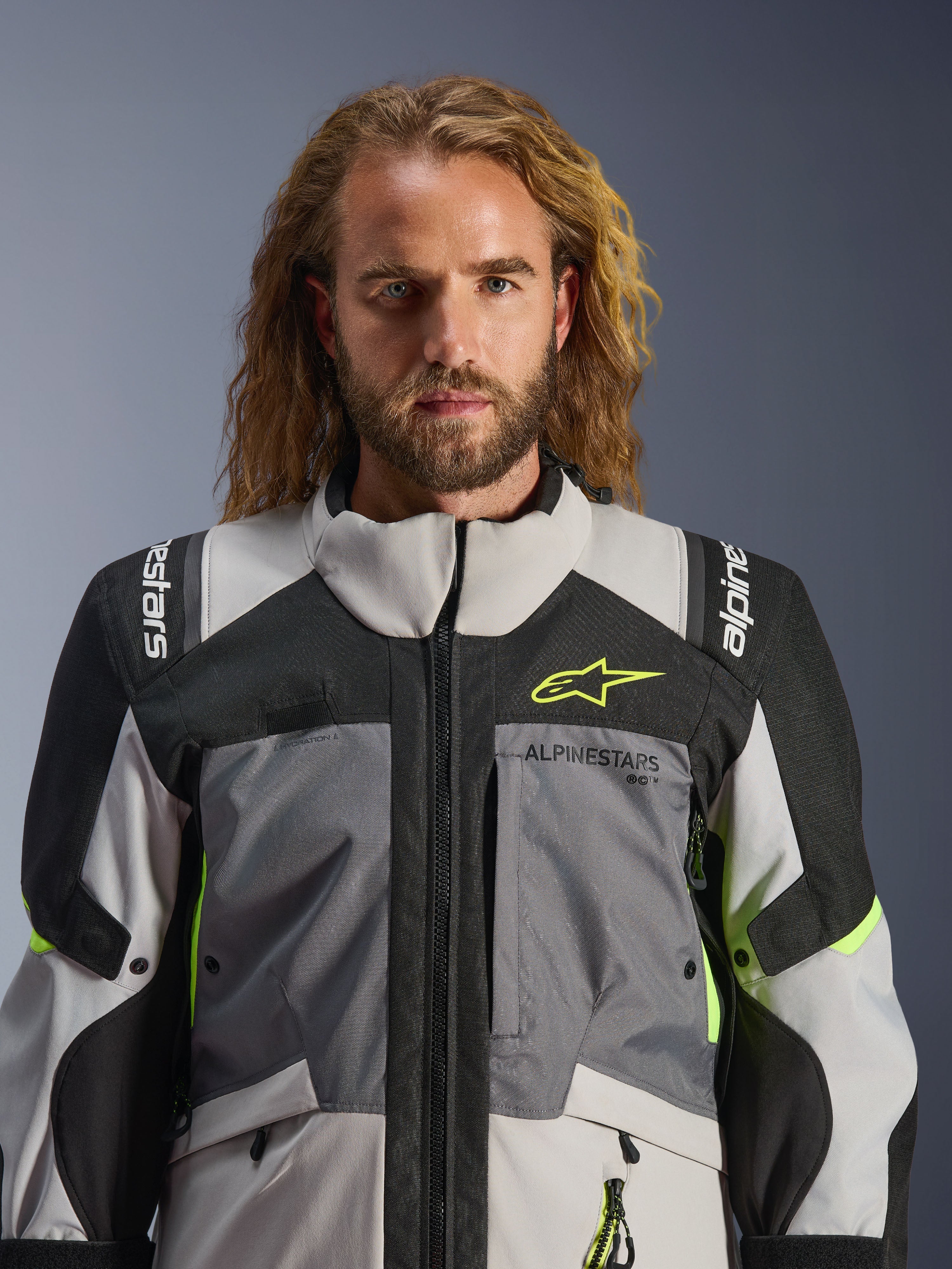 Giacca Alpinestars Andes V4 Drystar®, grigio scuro, grigio ghiaccio, nero e giallo fluo, indossata da un uomo con lunghi capelli mossi, caratterizzata da una costruzione tecnica multi-tessuto, loghi Alpinestars su petto e spalle, design a collo alto per la guida adventure
