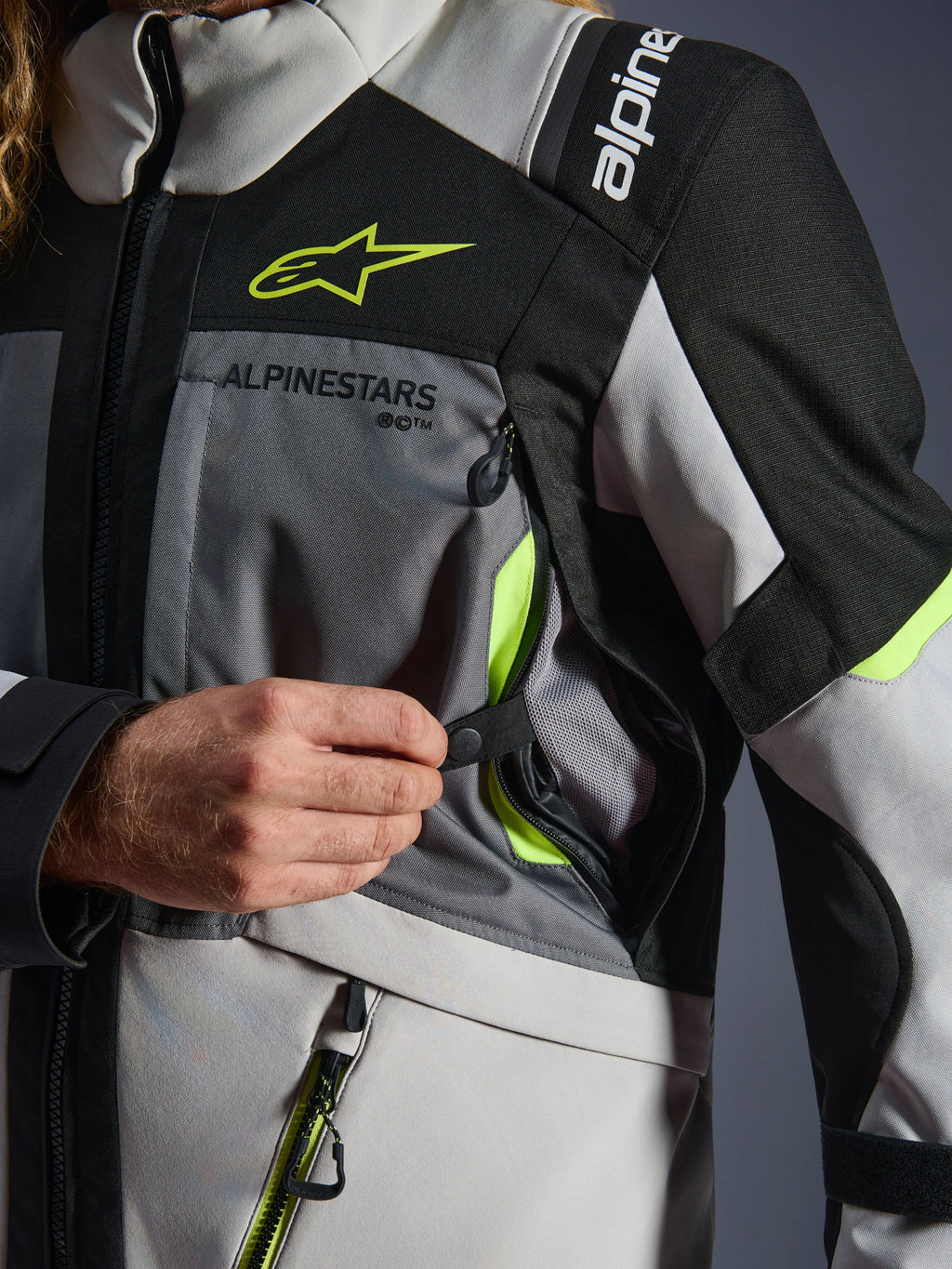 Giacca Alpinestars Andes V4 Drystar®, giacca da moto, Grigio Scuro/Grigio Ghiaccio/Nero/Giallo Fluo, dettaglio ravvicinato di una mano che apre la presa di ventilazione sul petto con accenti giallo neon e fodera in rete, costruzione in tessuto resistente con logo Alpinestars.
