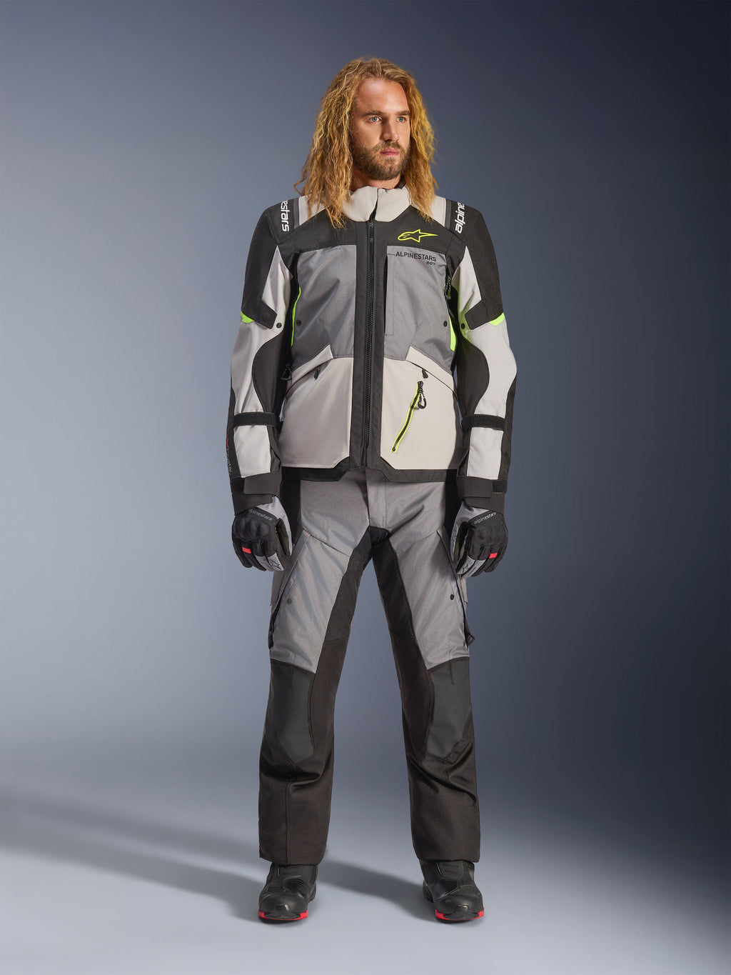Modello con lunghi capelli biondi che indossa la giacca Andes V4 Drystar® in Dark Gray, Ice Gray, Black e Yellow Fluo, abbinata a pantaloni da moto adventure coordinati e stivali neri, completo tecnico da guida per touring in ogni condizione meteorologica.