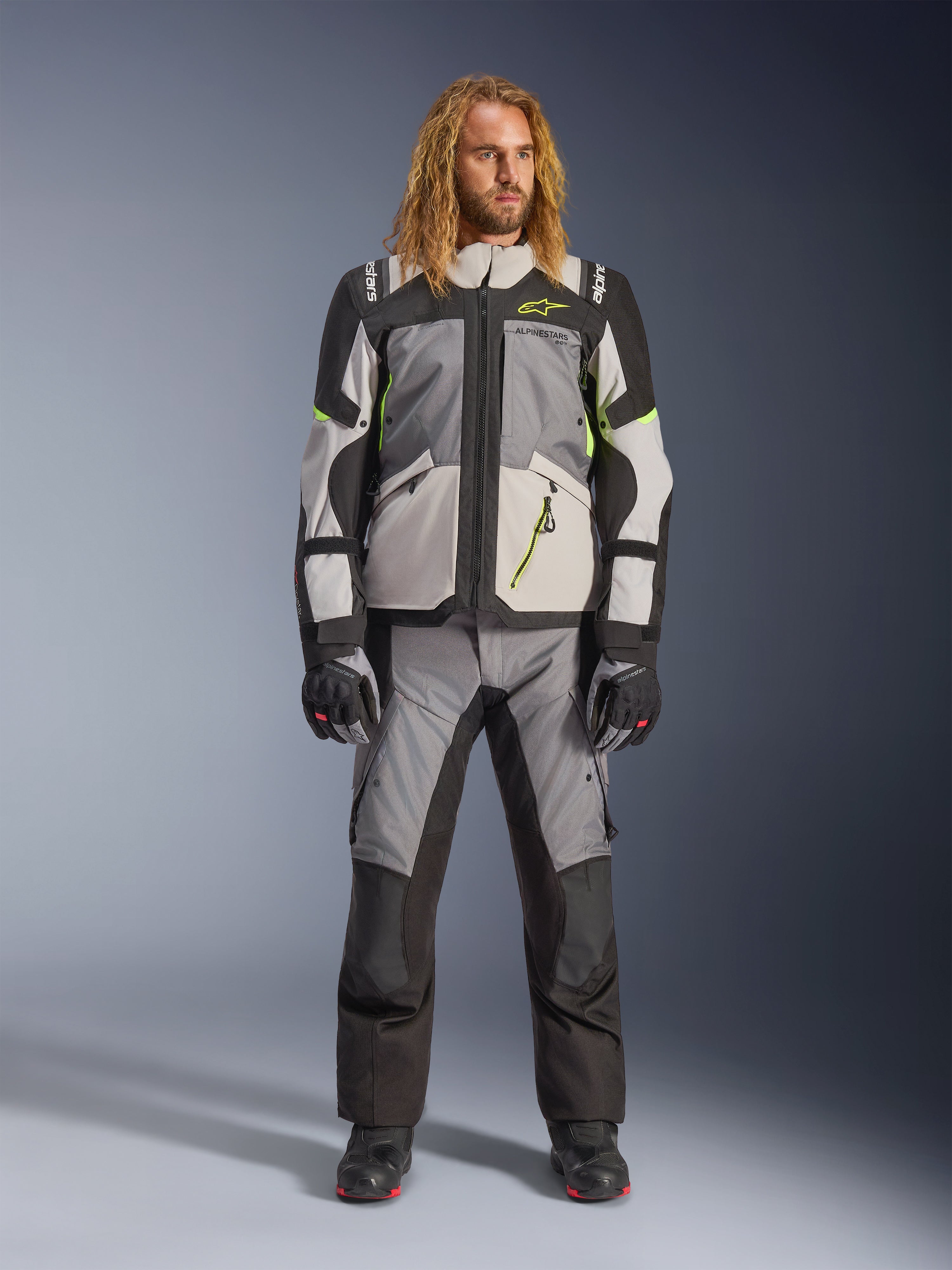 Modello con lunghi capelli biondi che indossa la giacca Andes V4 Drystar® in Dark Gray, Ice Gray, Black e Yellow Fluo, abbinata a pantaloni da moto adventure coordinati e stivali neri, completo tecnico da guida per touring in ogni condizione meteorologica.