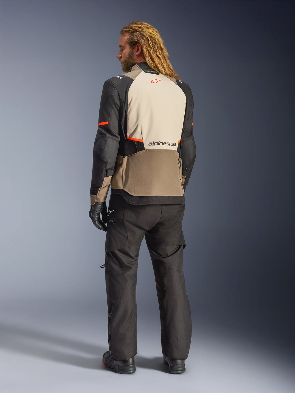 Giacca Alpinestars Andes V4 Drystar®, Walnut Mastic Black, vista posteriore su modello, pannelli in tessuto rinforzato color sabbia e nero con accenti arancioni, logo Alpinestars sulla schiena, indossata con pantaloni da moto adventure e guanti neri coordinati