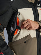 Dettaglio della giacca Alpinestars Andes V4 Drystar®, tessuto Walnut Mastic Black, primo piano di una mano che apre il pannello di ventilazione sul petto per rivelare l'interno in mesh arancione brillante, sistema di flusso d'aria diretto per la guida adventure
