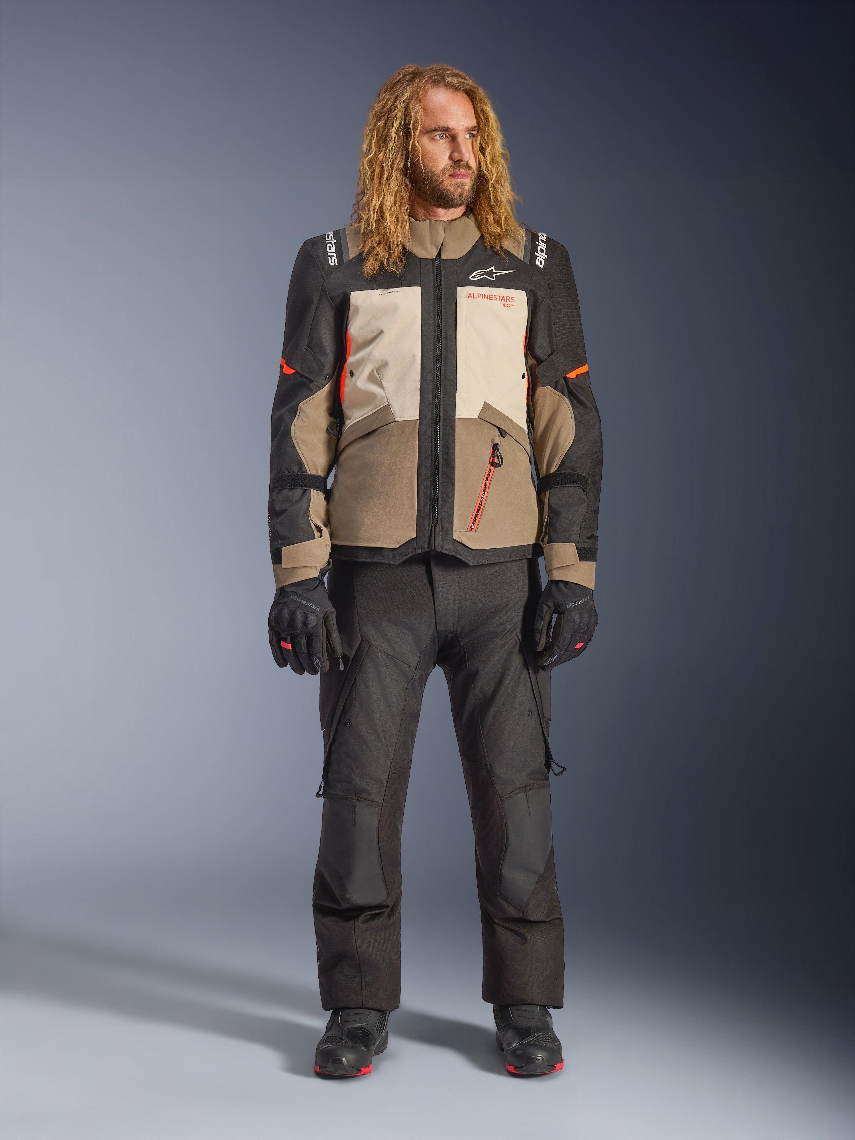 Giacca Alpinestars Andes V4 Drystar®, Walnut Mastic Black, indossata con pantaloni e guanti da moto neri, design adventure multi-pannello con accenti arancioni e marchio Alpinestars, abbigliamento tecnico in tessuto per ogni condizione atmosferica