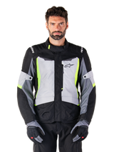 Giacca Alpinestars ST-1 Waterproof, giacca da moto adventure touring, Ice Gray, nero e giallo fluo, indossata da un uomo con guanti coordinati e pantaloni neri, con accenti giallo neon su petto e colletto, logo Alpinestars sul petto