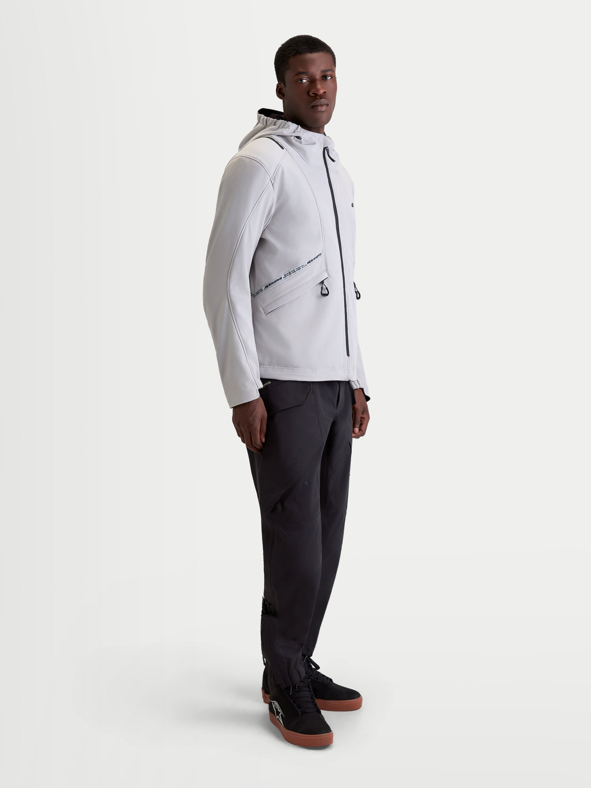 Alpinestars C 5 Drystar Giacca, giacca da moto urbana contemporanea da uomo in grigio chiaro e nero, vista di profilo laterale su un modello che indossa pantaloni scuri e scarpe da moto coordinati, struttura softshell impermeabile a 2 strati con cappuccio integrato