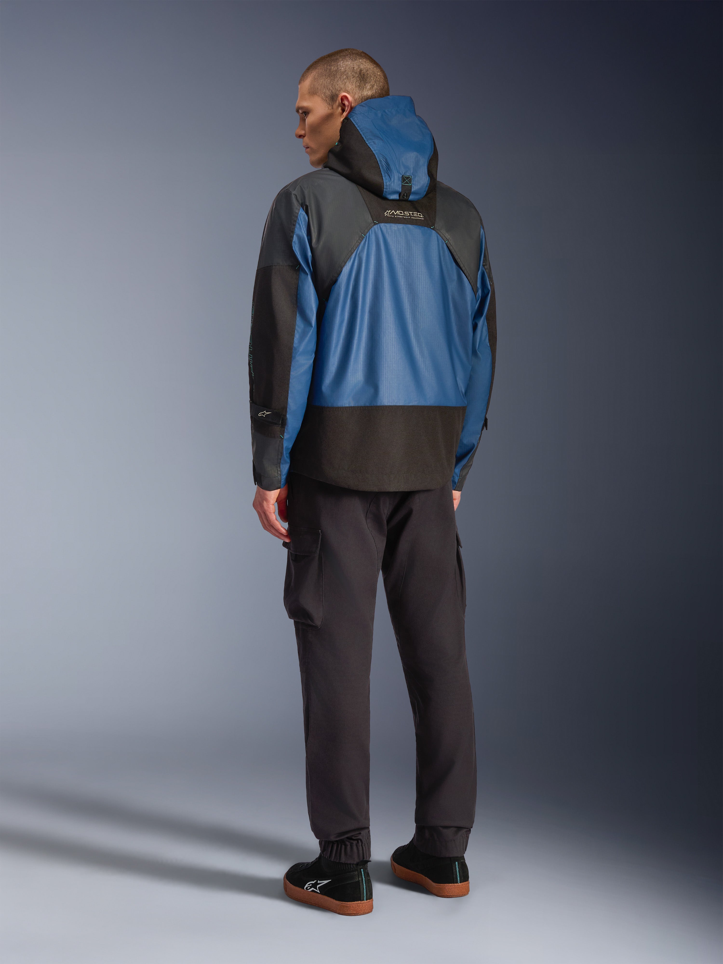 Vista posteriore di un modello che indossa la giacca Alpinestars Mospher Waterproof in nero e Dark Petrol, caratterizzata da un design con cappuccio e costruzione tattica in ripstop, abbinata a pantaloni cargo neri e sneaker.