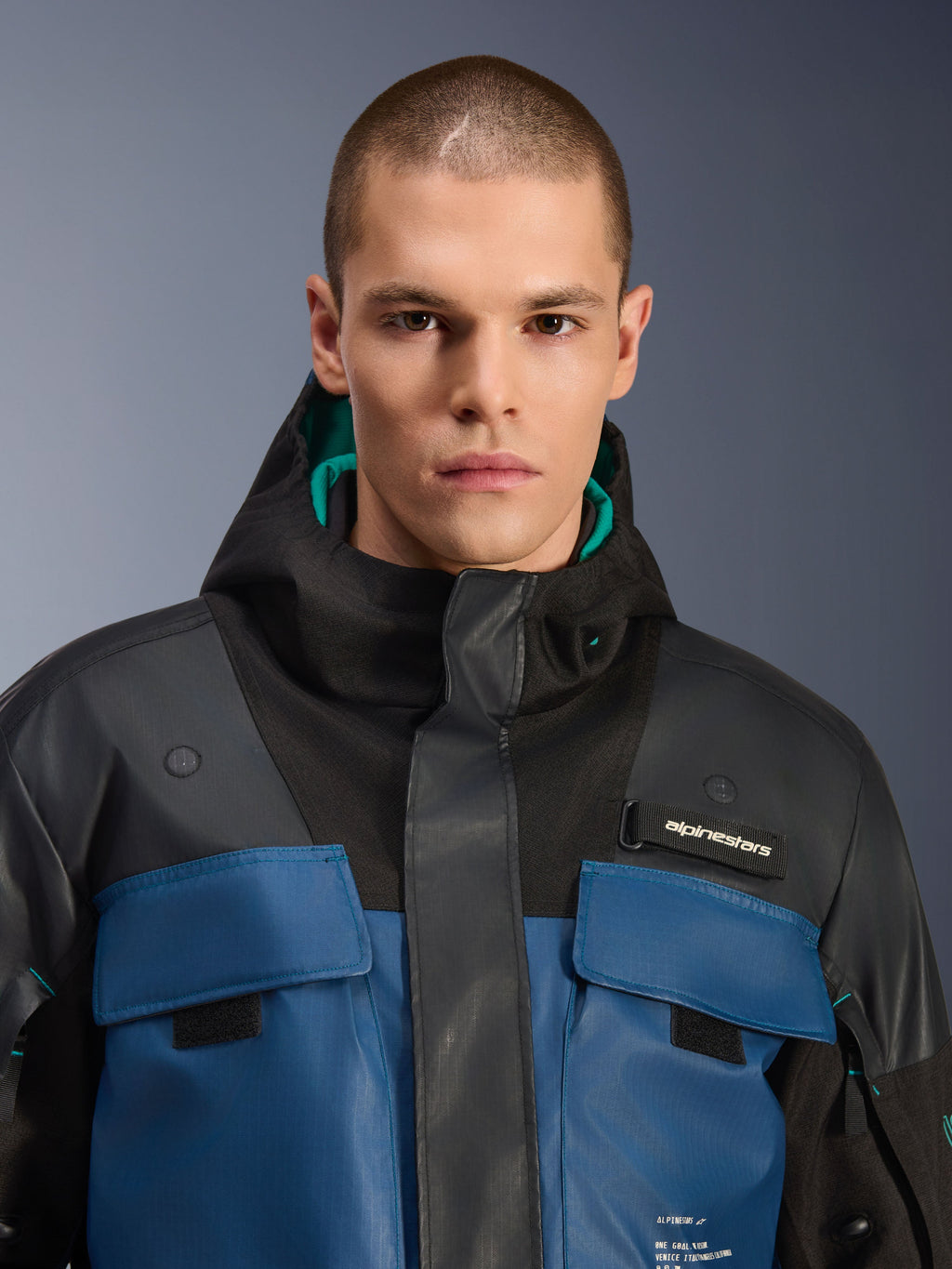 Giacca Alpinestars Mospher Waterproof, nero e Dark Petrol, primo piano di un modello maschile che indossa il parka d'ispirazione utility, con tasche sul petto blu scuro, cappuccio nero con fodera color ottanio e logo Alpinestars sul petto.