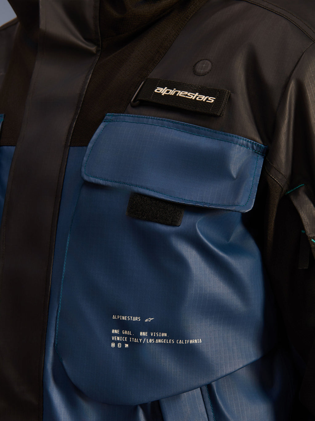 Giacca Alpinestars Mospher Waterproof, dettaglio ravvicinato della tasca sul petto, colore Nero e Dark Petrol, robusto tessuto ripstop con stampa 'One Goal. One Vision.', design funzionale con tasche e linguetta con logo Alpinestars
