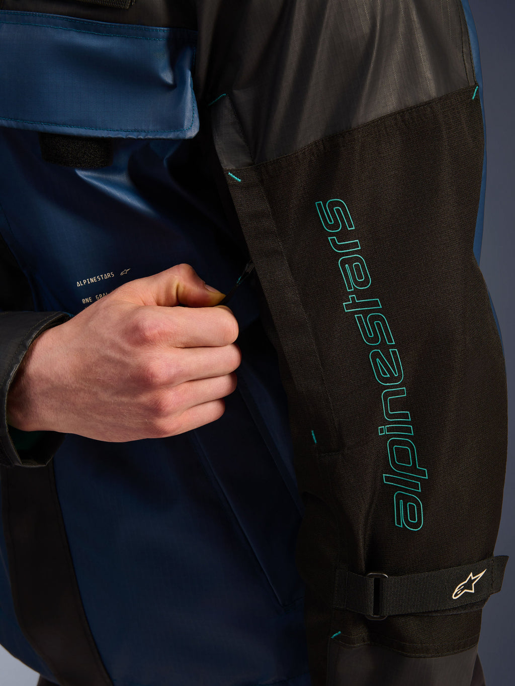 Giacca Alpinestars Mospher WP in Nero Dark Petrol, dettaglio della manica con branding Alpinestars color petrolio, una mano che regola la cerniera di ventilazione, tessuto ripstop resistente e cinturino regolabile sul braccio