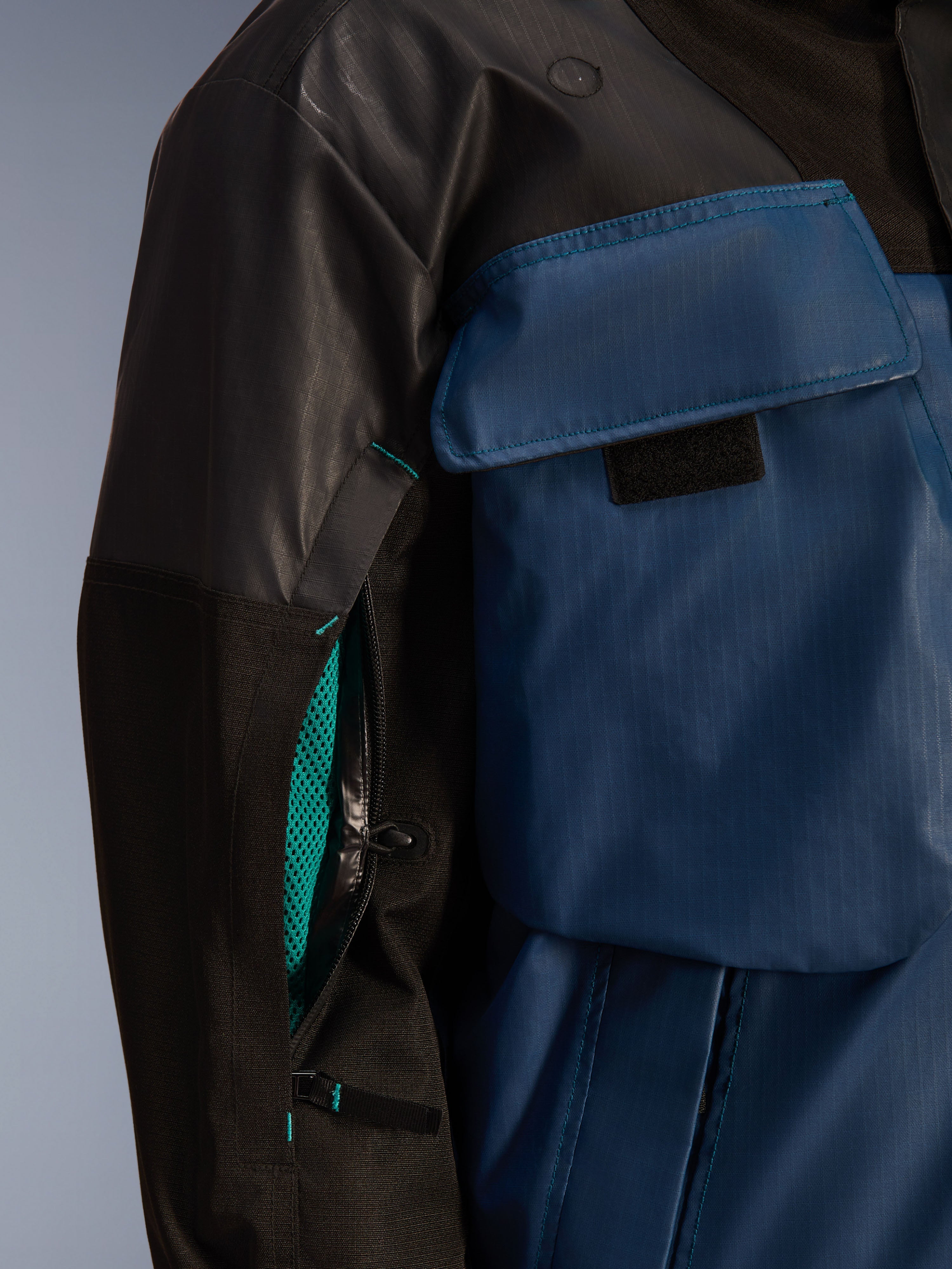 Primo piano della Giacca Alpinestars Mospher WP in Nero e Dark Petrol, con una cerniera di ventilazione aperta sulla manica con fodera in rete verde acqua brillante, grande tasca sul petto e materiale in tessuto ripstop resistente.