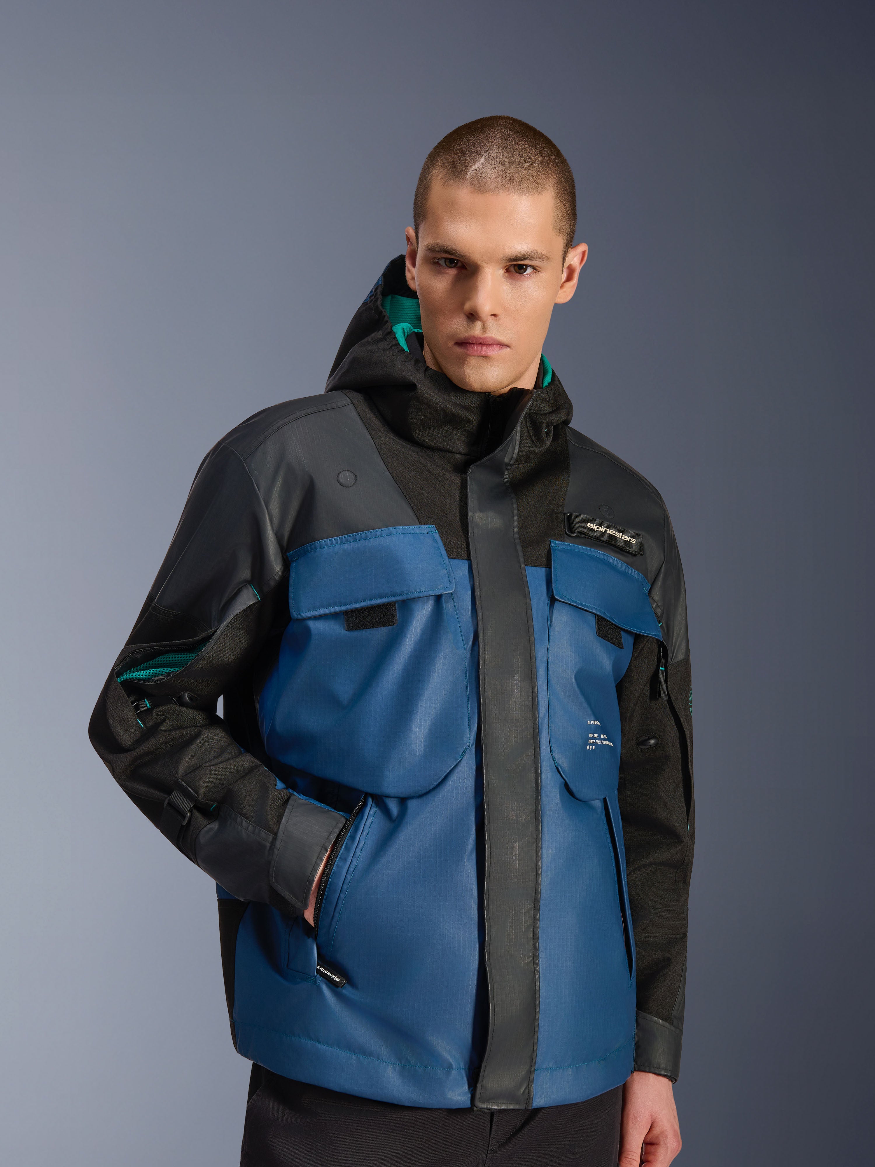 Modello che indossa la giacca impermeabile Alpinestars Mospher in nero e Dark Petrol, con ampie tasche utility sul petto, design con cappuccio e fodera verde acqua, in tessuto tecnico ripstop per il commuting urbano in moto.