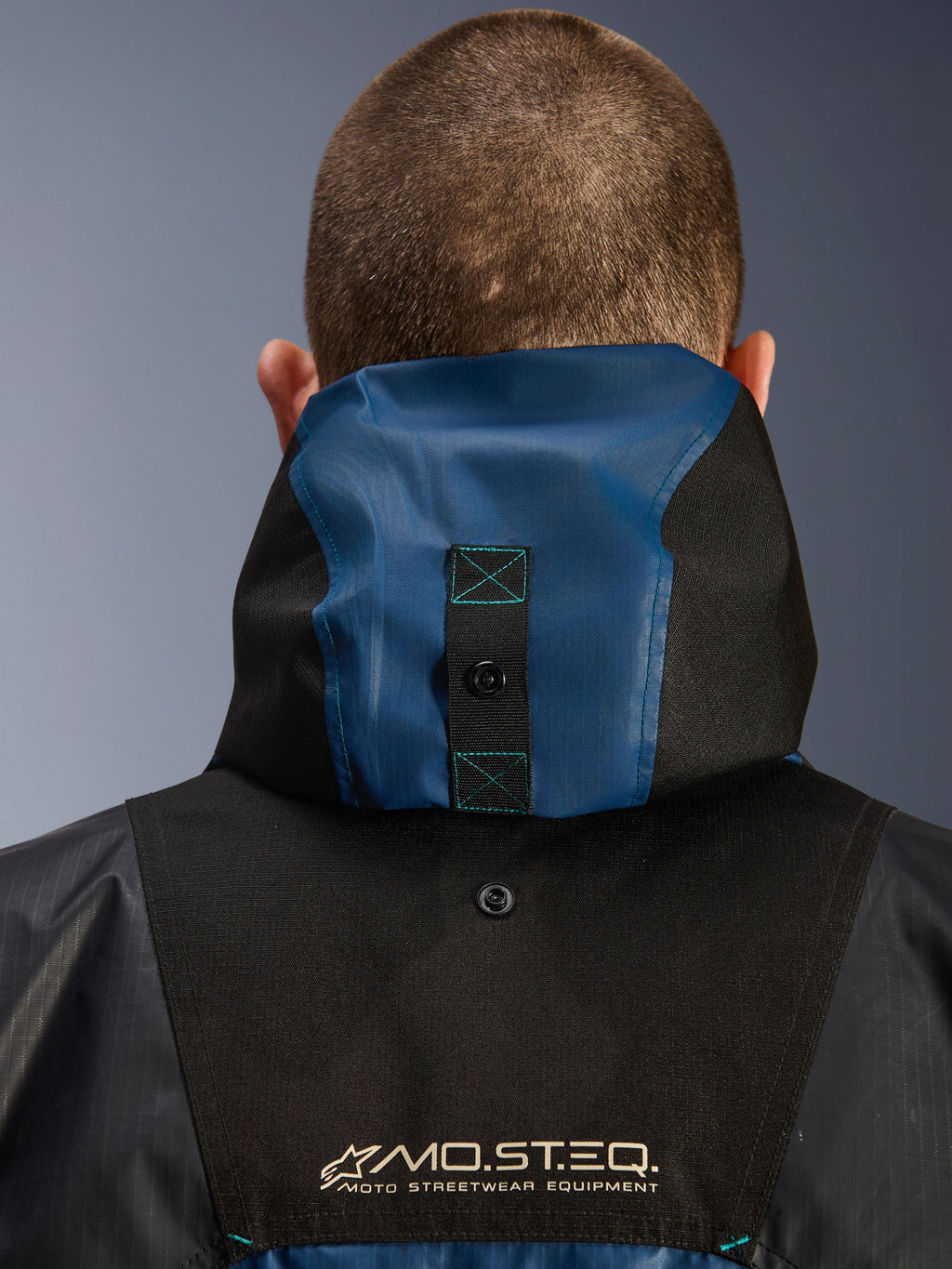 Alpinestars Mospher Waterproof Jacket, dettaglio della vista posteriore con sistema di ancoraggio del cappuccio, nero e blu petrolio scuro, logo MO.ST.EQ. Moto Streetwear Equipment, materiale in tessuto ripstop resistente, abbigliamento tecnico da moto per pendolari