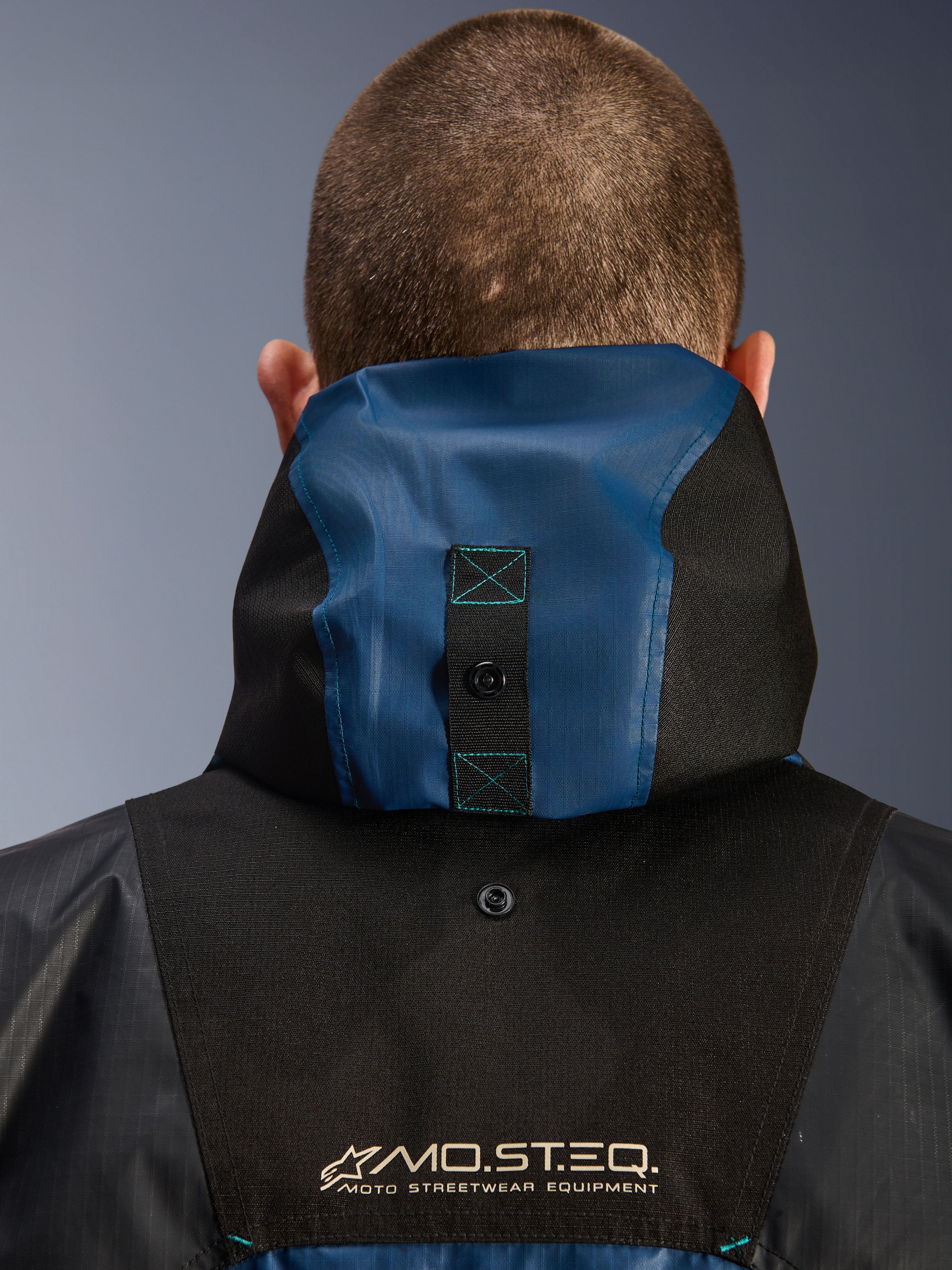 Alpinestars Mospher Waterproof Jacket, dettaglio della vista posteriore con sistema di ancoraggio del cappuccio, nero e blu petrolio scuro, logo MO.ST.EQ. Moto Streetwear Equipment, materiale in tessuto ripstop resistente, abbigliamento tecnico da moto per pendolari