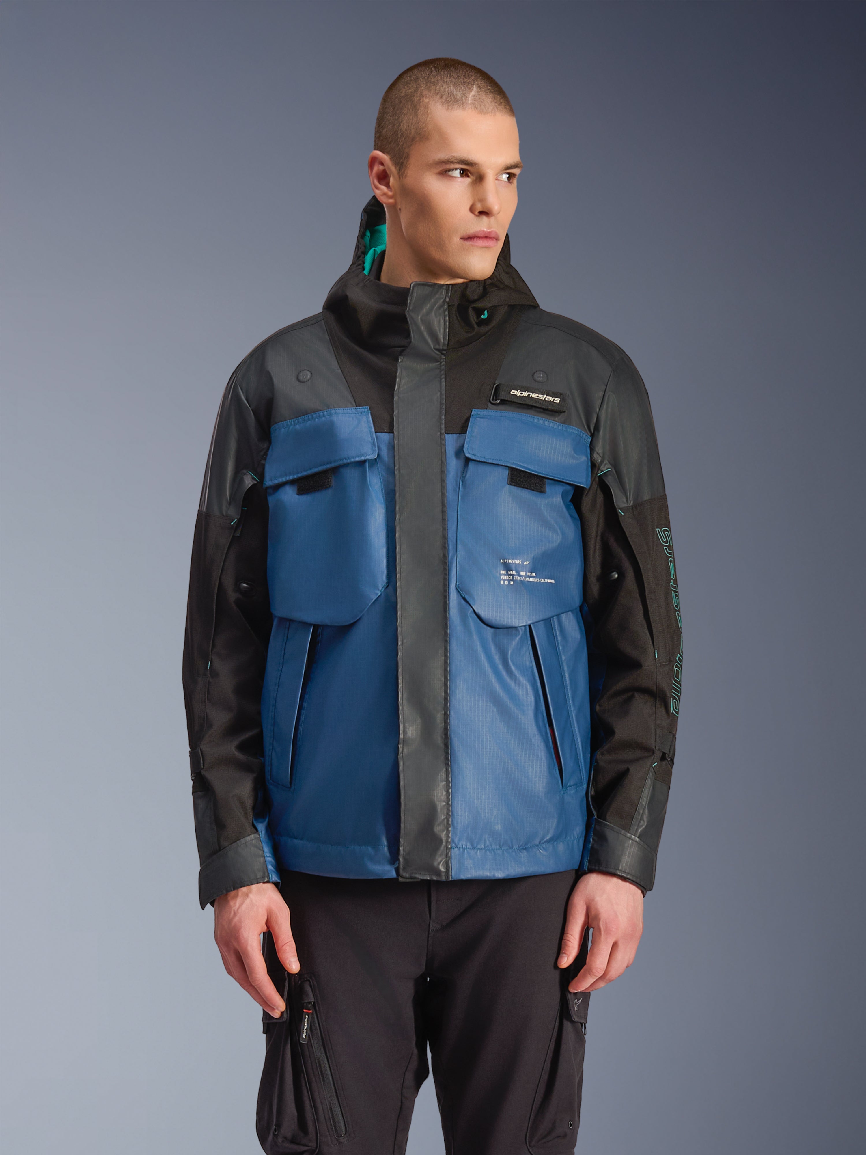Alpinestars Mospher Waterproof Jacket, parka moto urban, nero e blu petrolio scuro, con grandi tasche tattiche sul petto, collo alto con cappuccio, trama tecnica ripstop, vista frontale su modello