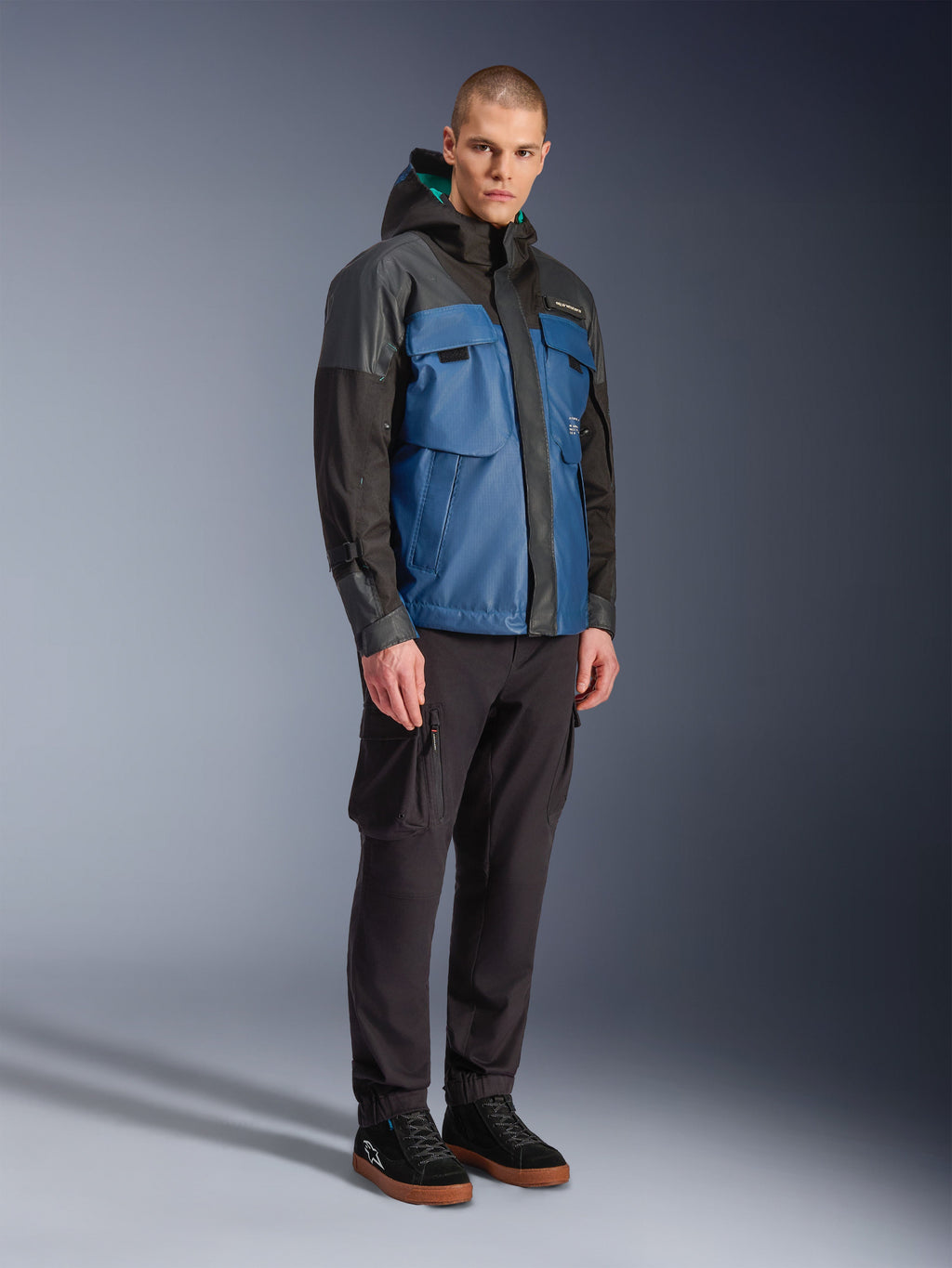 Giacca Alpinestars Mospher Waterproof, nero e blu petrolio scuro, indossata con pantaloni cargo neri e sneakers nere, stile urban commuter con tasche utility e cappuccio