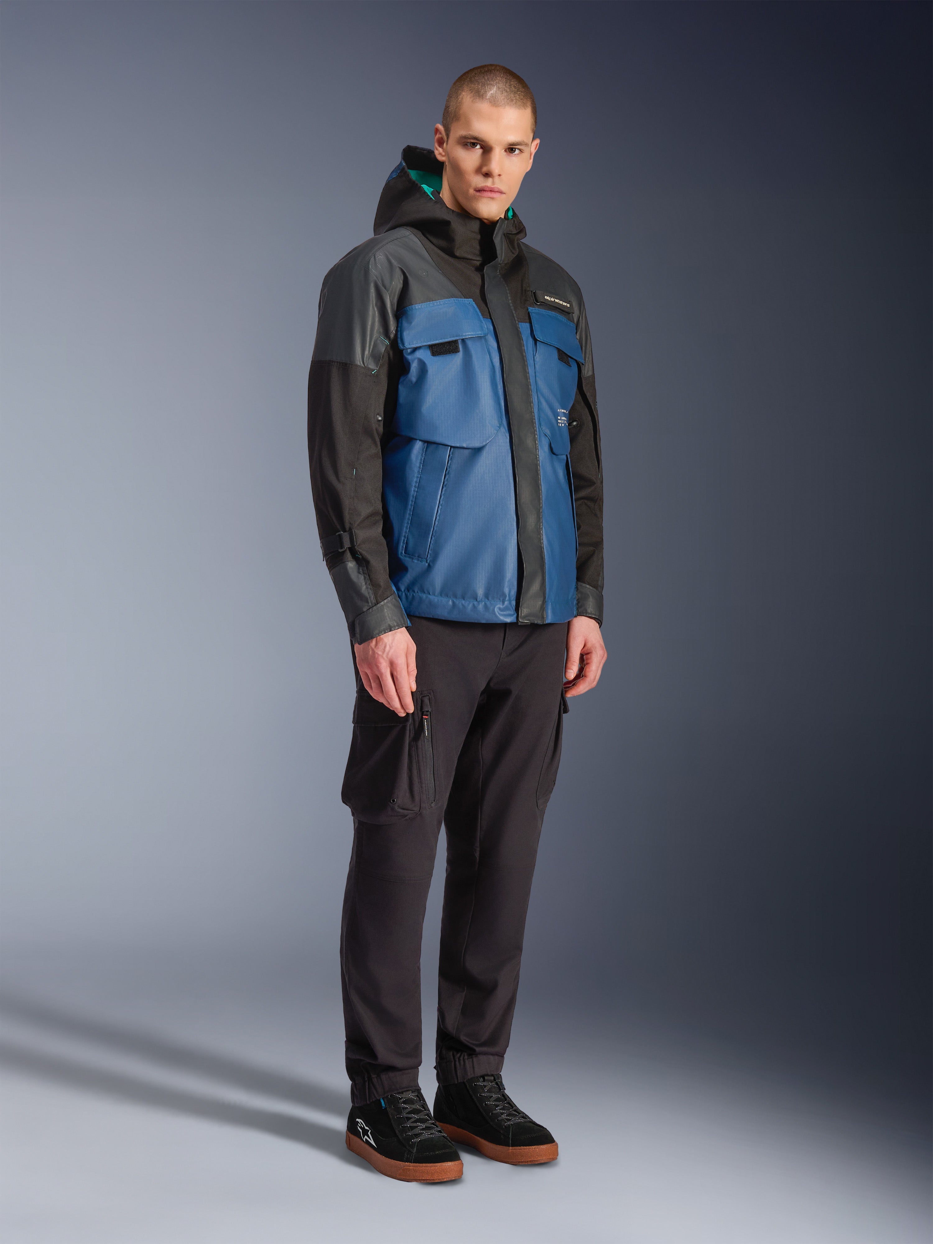 Giacca Alpinestars Mospher Waterproof, nero e blu petrolio scuro, indossata con pantaloni cargo neri e sneakers nere, stile urban commuter con tasche utility e cappuccio