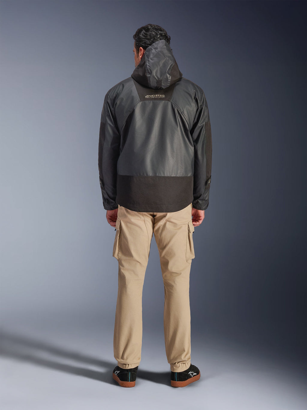 Alpinestars Giacca Mospher Waterproof, parka da moto nero visto dal retro, con logo MO.ST.EQ sulla parte superiore della schiena e struttura in tessuto ripstop, indossata con pantaloni cargo color sabbia e sneaker nere, stile urban commuter.