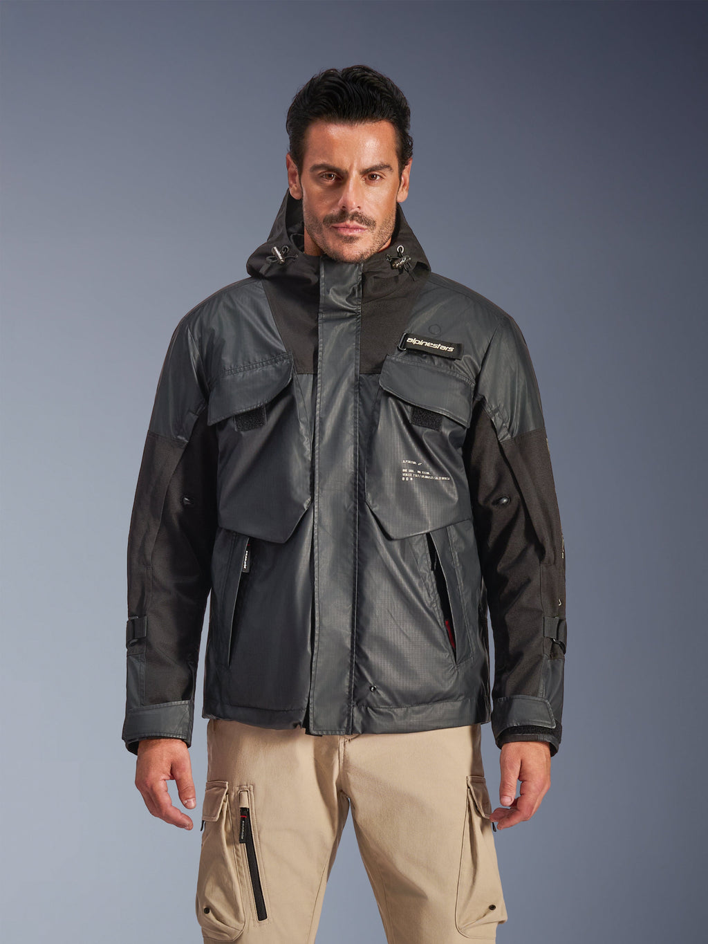 Alpinestars Mospher Waterproof Jacket, parka da moto urban da uomo, tessuto ripstop nero e grigio scuro, design con cappuccio e ampie tasche multiuso, indossata da un modello con pantaloni cargo kaki per un look tattico.