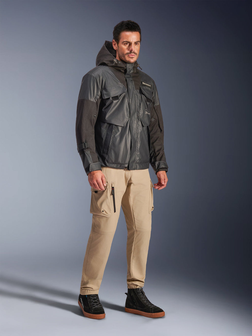 Modello che indossa la giacca Alpinestars Mospher Waterproof in nero, parka impermeabile d'ispirazione utility con design multitasche e cappuccio, abbinato a pantaloni cargo color sabbia e sneaker alte nere, abbigliamento moto urban per pendolari.