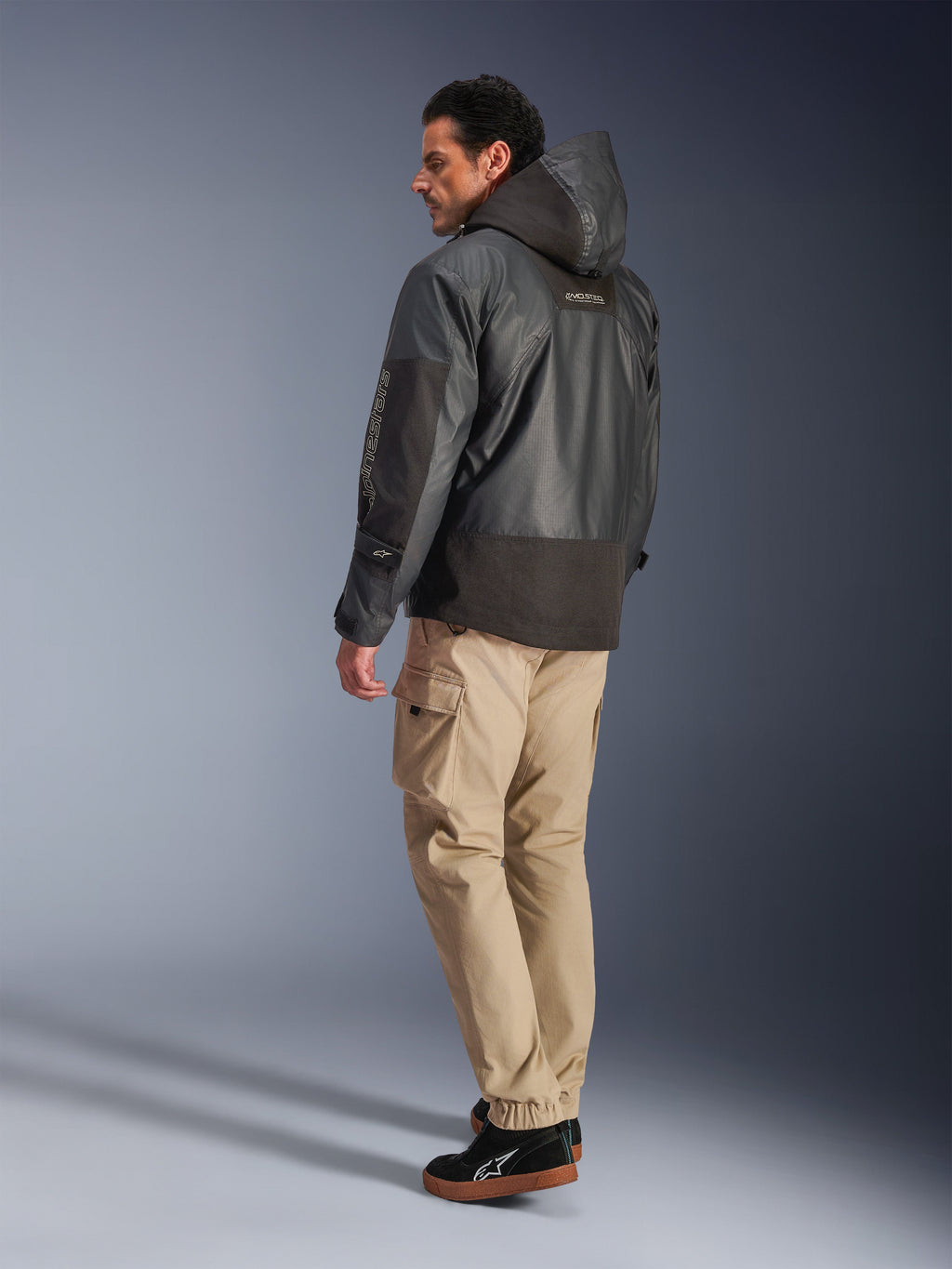Uomo che indossa la giacca impermeabile Alpinestars Mospher WP in nero, vista posteriore e di profilo laterale, abbinata a pantaloni cargo color sabbia e sneakers nere. Design parka con cappuccio in tessuto ripstop, marchio Alpinestars sulla manica e stile urban moderno per motociclisti.