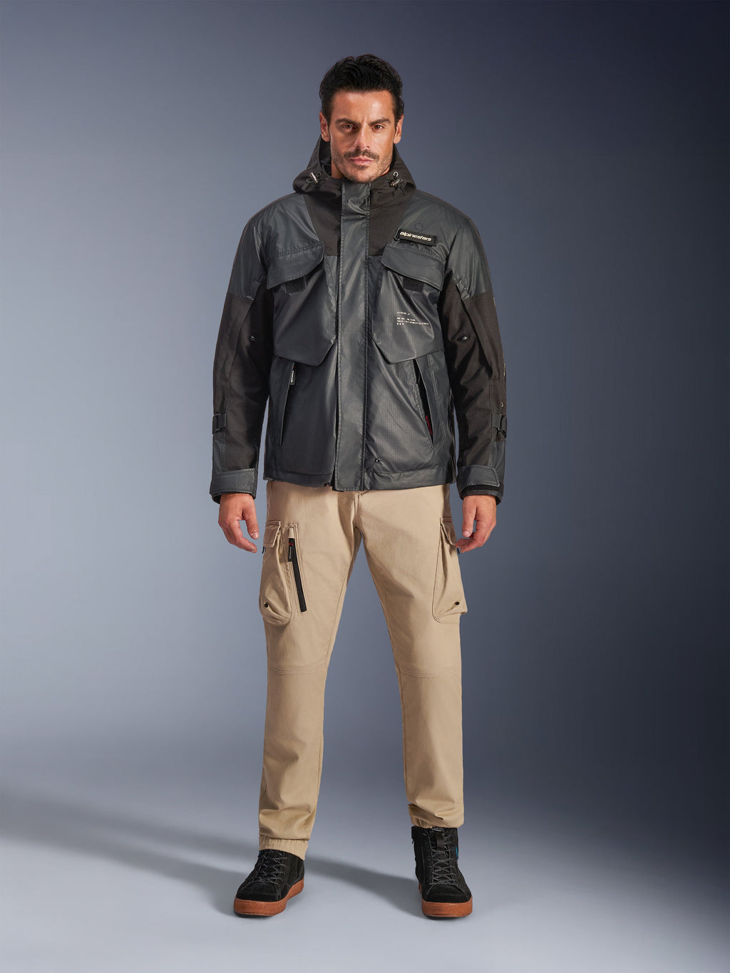 Modello che indossa la giacca impermeabile Alpinestars Mospher in nero, abbigliamento da moto urban contemporary, design tattico utility con molteplici tasche funzionali e cappuccio, abbinata a pantaloni cargo color sabbia e scarpe nere, ritratto in studio a figura intera.
