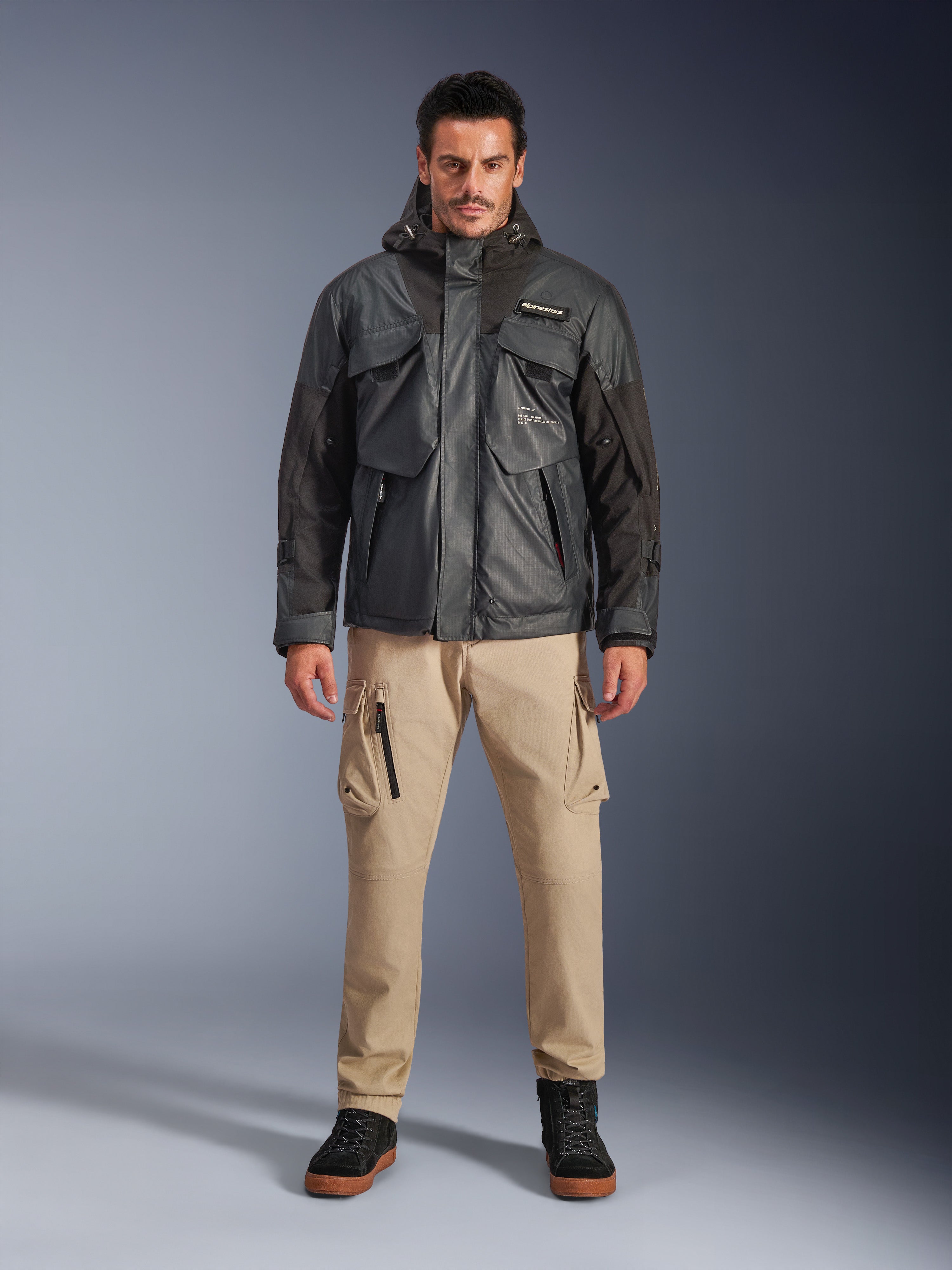 Modello che indossa la giacca impermeabile Alpinestars Mospher in nero, abbigliamento da moto urban contemporary, design tattico utility con molteplici tasche funzionali e cappuccio, abbinata a pantaloni cargo color sabbia e scarpe nere, ritratto in studio a figura intera.