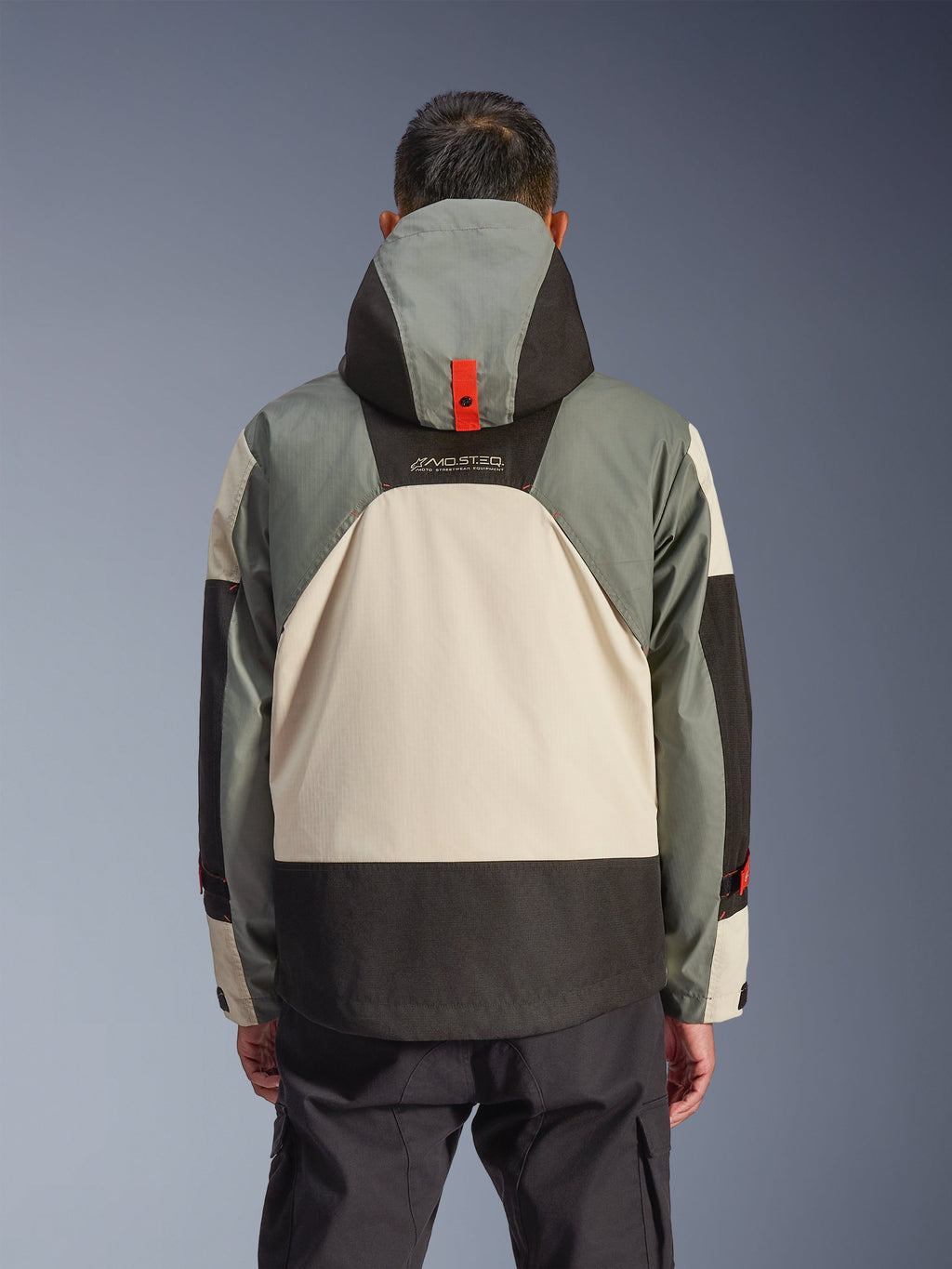 Vista posteriore della giacca Alpinestars Mospher Waterproof in Fur Gray Black Ice, caratterizzata da un design a pannelli grigio multi-tonale, crema e nero, silhouette parka con cappuccio e dettaglio di ancoraggio rosso, marchio 'MO.ST.EQ.', abbinata a pantaloni cargo da moto neri.