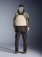 Vista posteriore di un pilota che indossa la giacca impermeabile Alpinestars Mospher in Fur Gray Black Ice, parka d'ispirazione tattica con design a blocchi di colore grigio, nero e crema, cappuccio con dettaglio arancione, abbinata a pantaloni cargo neri