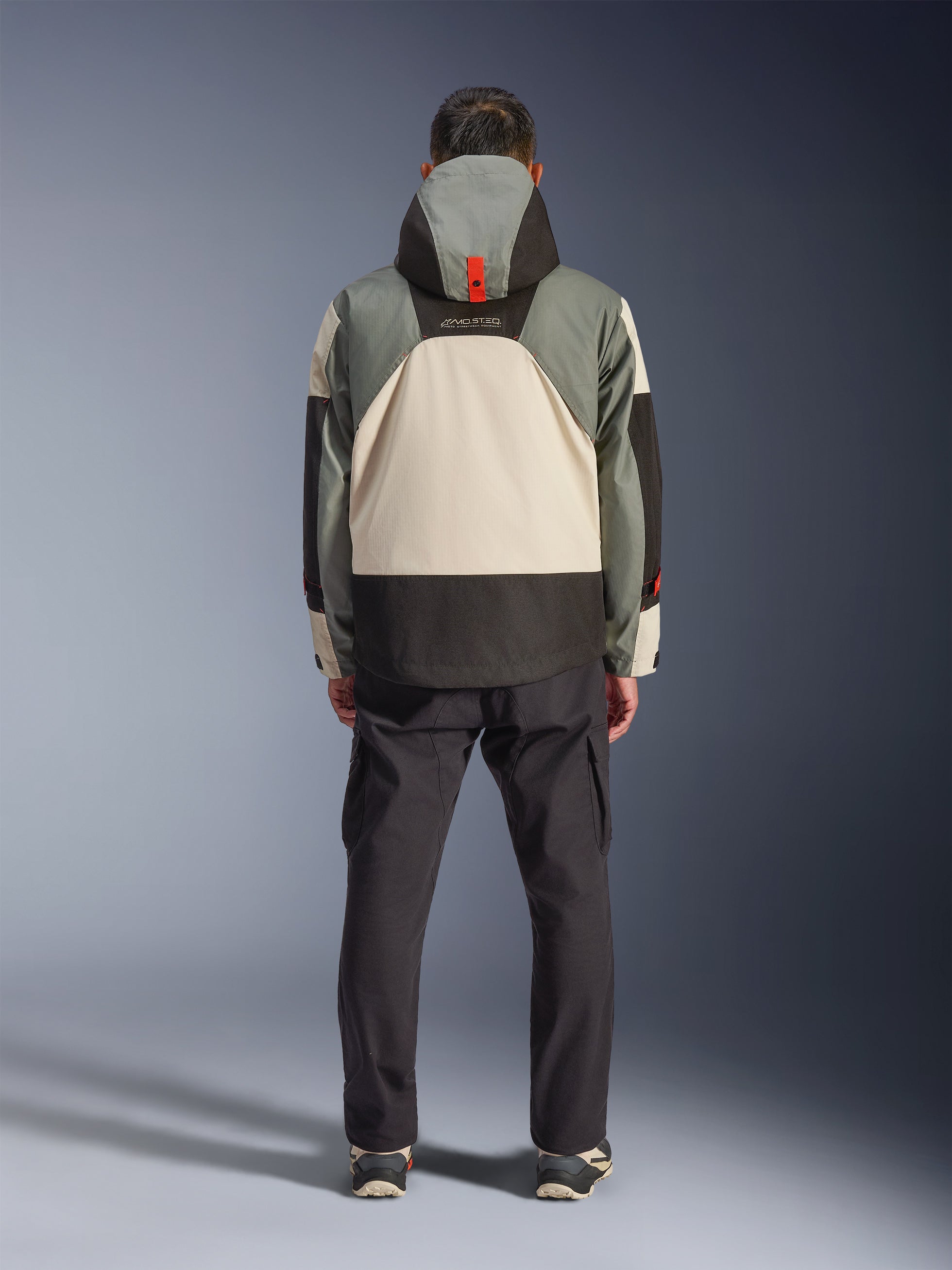 Vista posteriore di un pilota che indossa la giacca impermeabile Alpinestars Mospher in Fur Gray Black Ice, parka d'ispirazione tattica con design a blocchi di colore grigio, nero e crema, cappuccio con dettaglio arancione, abbinata a pantaloni cargo neri