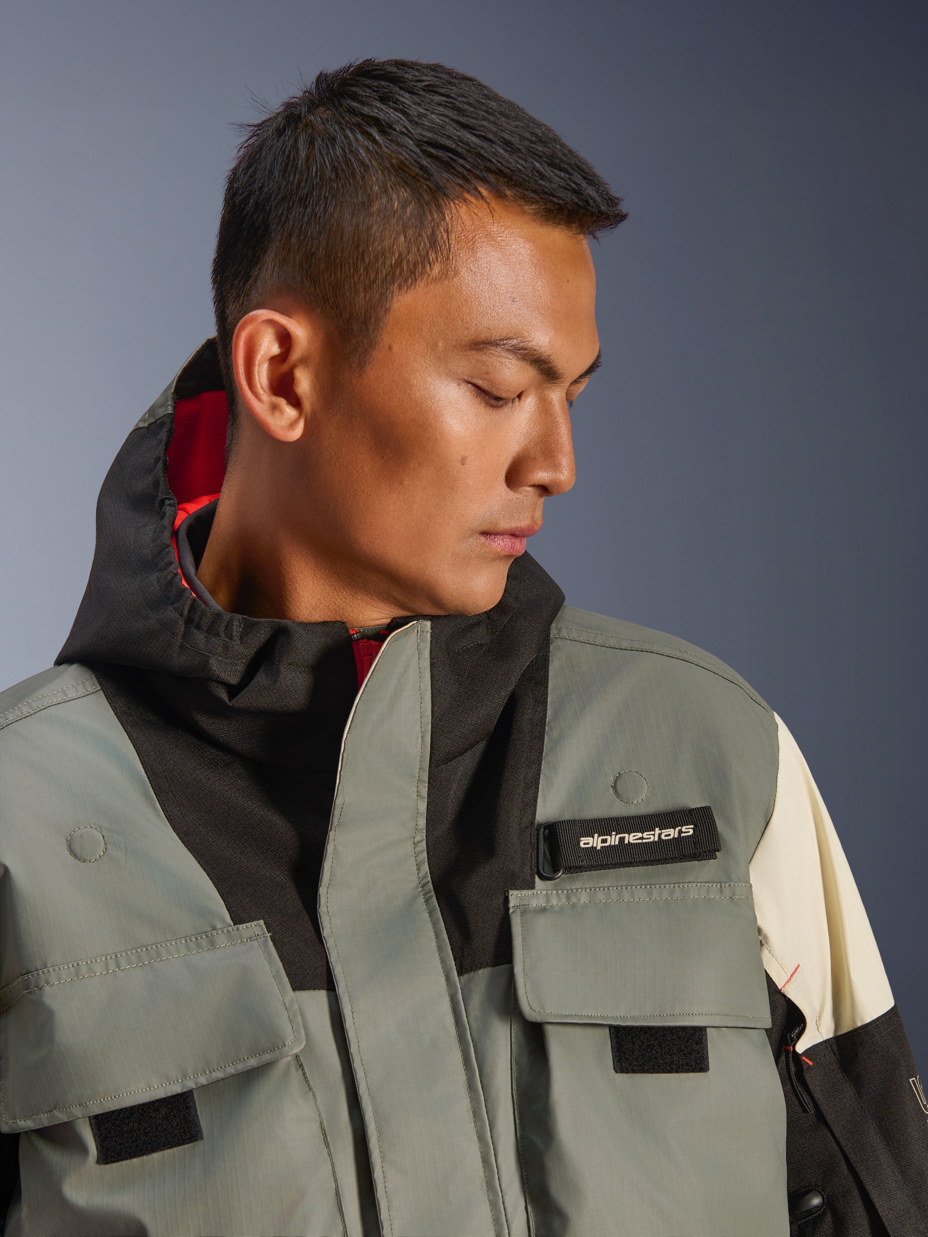 Giacca Alpinestars Mospher WP, parka da moto impermeabile, Fur Gray Black Ice, primo piano del modello che indossa la giacca con cappuccio e tasche utility, linguetta nera con logo Alpinestars e design tattico con fodera interna rossa