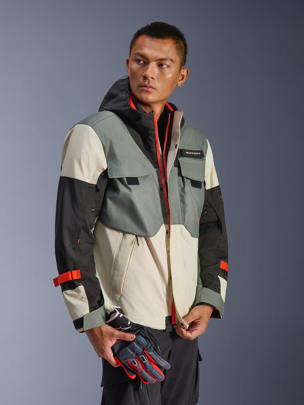 Giacca Alpinestars Mospher Waterproof in Fur Gray Black Ice, parka in tessuto multi-pannello grigio, nero e bianco sporco, dettagli rosso vivace su cerniere e cinturini, design d'ispirazione tattica con cappuccio e tasche multiuso, indossata da un modello con guanti da moto