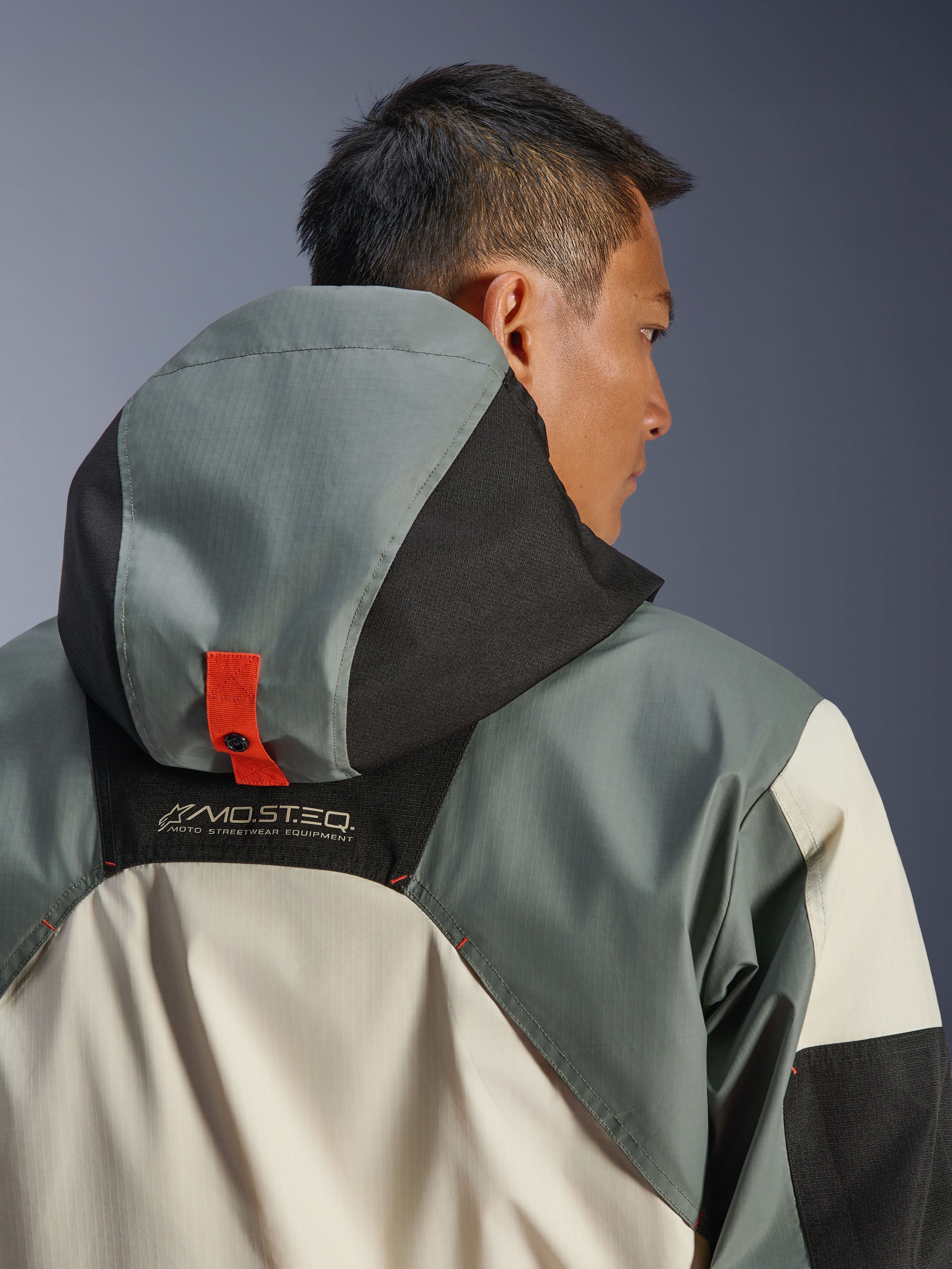 Giacca impermeabile Alpinestars Mospher, Fur Gray Black Ice, vista posteriore ravvicinata del sistema di ancoraggio del cappuccio con linguetta arancione e bottone a pressione, marchio MO.ST.EQ. Moto Streetwear Equipment, costruzione in resistente tessuto ripstop