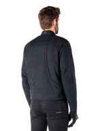 Giacca Alpinestars Clayton WR in nero, vista posteriore di tre quarti su un modello maschile, giacca da moto urbana con imbottitura trapuntata su spalle e gomiti, linee di cucitura tecniche e logo riflettente sul colletto, abbinata a pantaloni e guanti neri.
