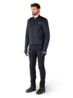 Pilota che indossa la giacca Alpinestars Clayton WR, giacca da moto urban nera, design slim-fit con dettagli trapuntati sulle spalle e finitura opaca, abbinata a pantaloni tecnici da guida e guanti neri.